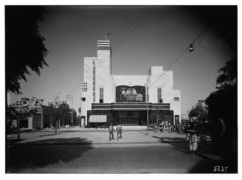 Alhambra Theater in Yafa, 1937. Image depicts the building facade with a Palestinian flag flying from its roof, a film poster with Arabic writing, and people walking near the building.