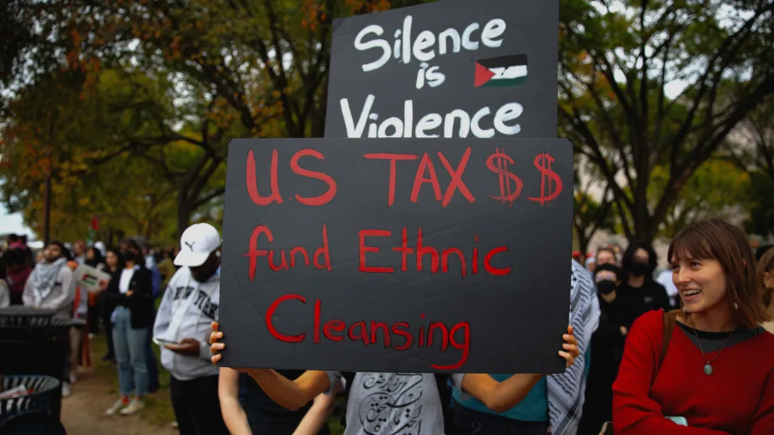 Protestor carries sign that says "Silence is Violence - US Tax $$ Fund Ethnic Cleansing" 