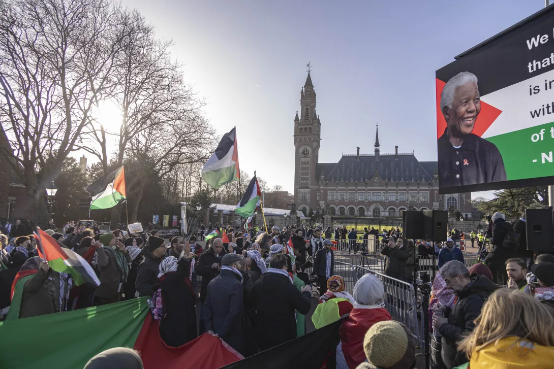 A crowd rallies carrying Palestinian flags and wearing kuffiyeh outside of the International Court of Justice in the Hague during proceedings initiated by South Africa against Israel for the crime of genocide. A large sign displays an image of Nelson Mandela.