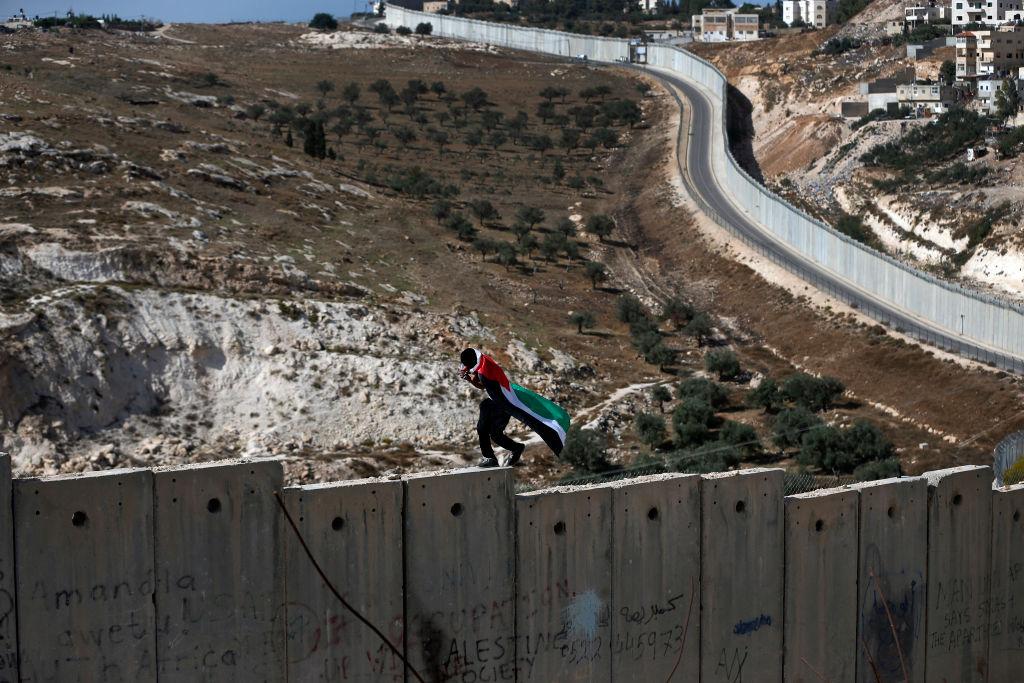 A man draped in the Palestinian flag walks on top of Israel's apartheid wall