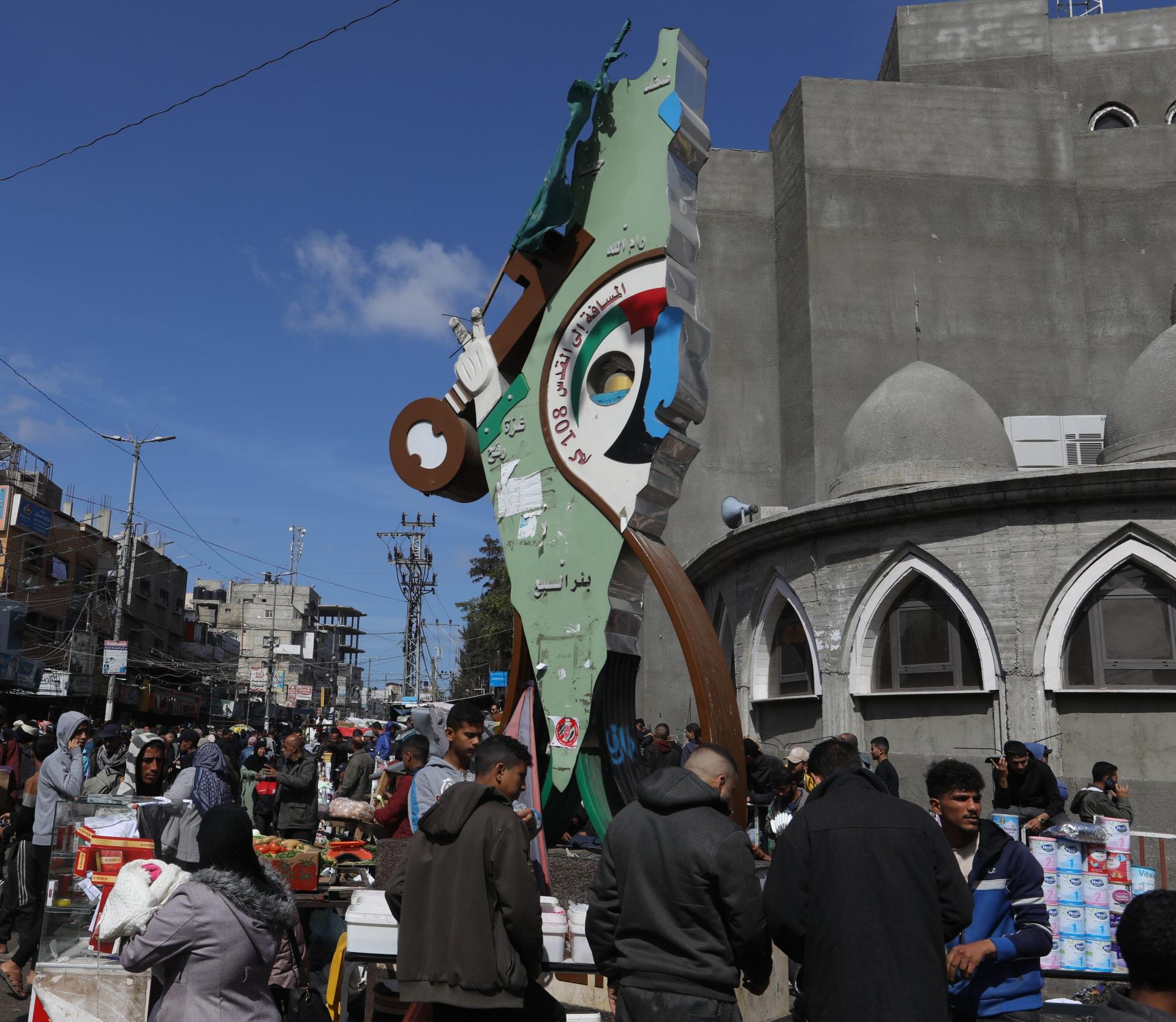 Displaced Palestinians shop at a market in Rafah near a statue marking "Return (`Awda) Square," displaying a map of historic Palestine, a key symbolizing the right of return, that marks the 108-kilometer distance from the landmark in Rafah to Jerusalem.