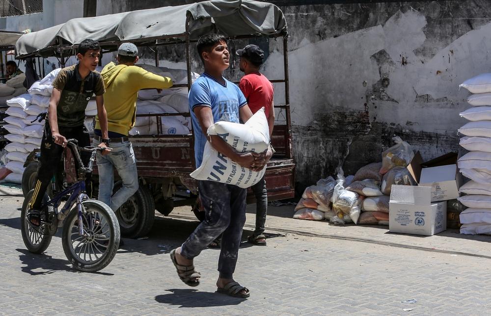 Palestinians collect food aid from an UNRWA distribution center in Rafah, a youth carries a flour sack.