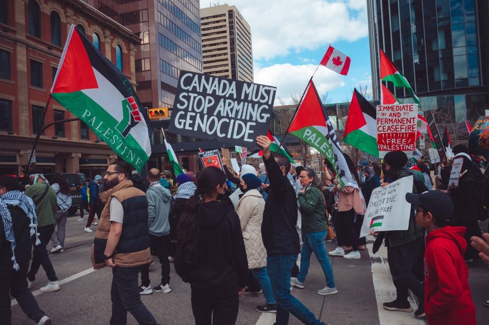 Protestors in Calgary, Canada wave Palestinian flags, a Canadian flag, and signs demanding a ceasefire in Gaza. One prominent sign reads "CANADA MUST STOP ARMING GENOCIDE."