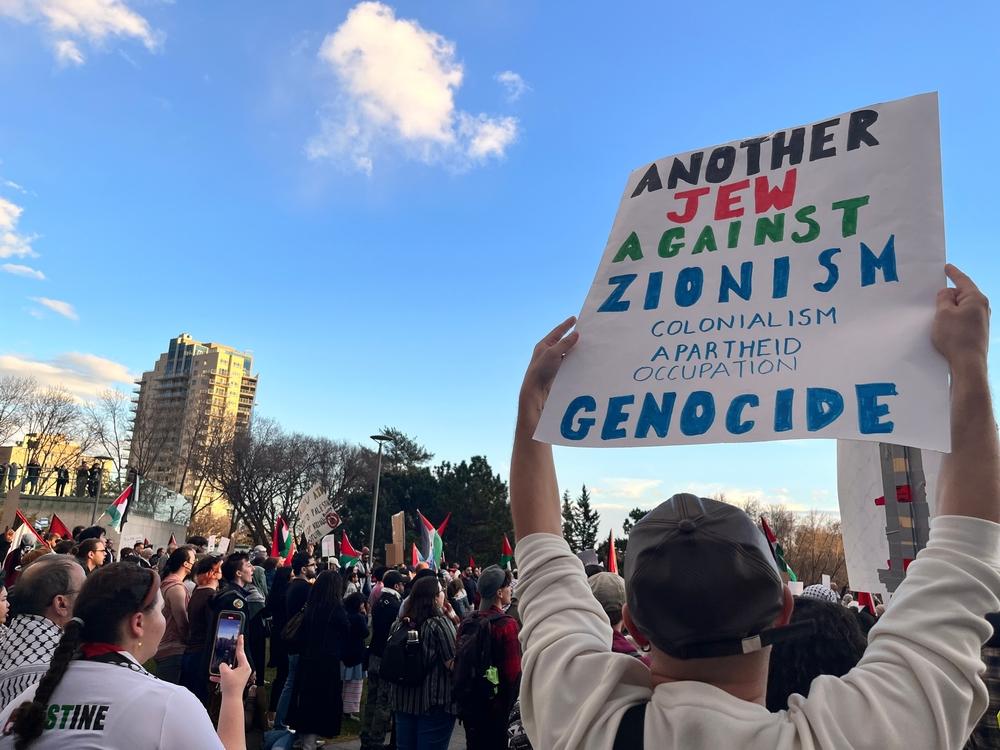 Protestor carries a sign that says "Another Jew Against Zionism, Colonialism, Apartheid, Occupation, Genocide."