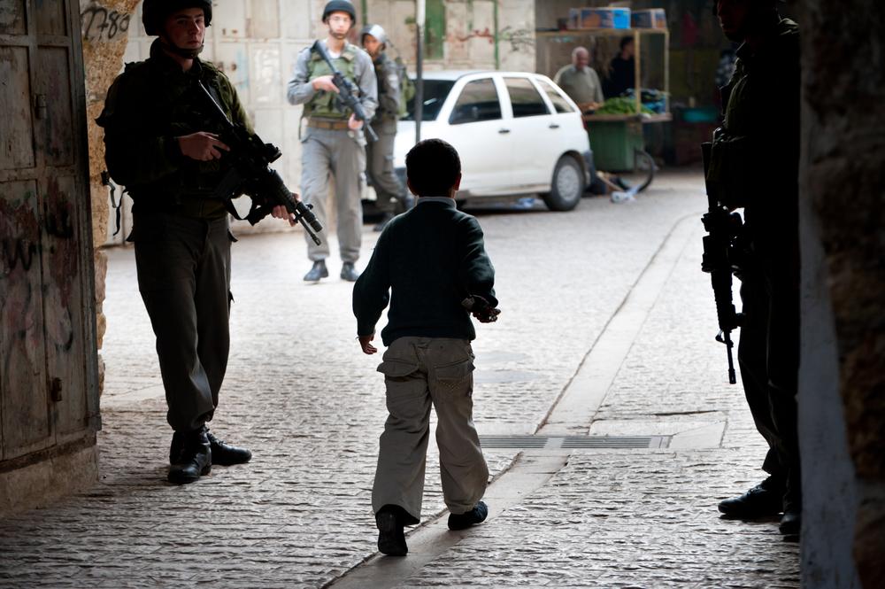 A Palestinian boy walks on a Hebron street surrounded by three Israeli soldiers carrying arms.