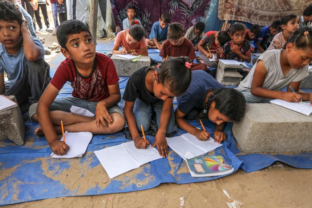 Displaced Palestinian children in a Deir al-Balah tent resume their education after two years of distruption due to Israel's genocide. They are writing in workbooks and sitting on the ground.