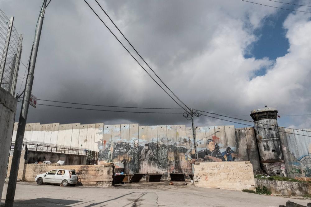 Image of Israel's apartheid wall with graffiti of the Palestinian struggle, including Palestinian with a slingshot and a blindfolded Palestinian being arrested by Israeli soldiers.