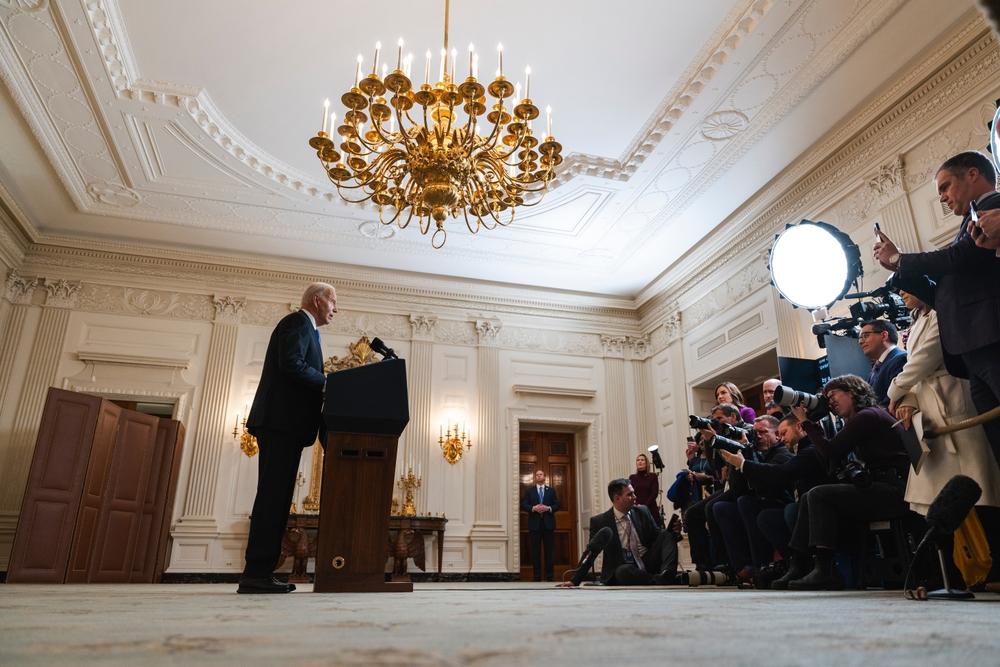 President Biden is pictured speaking at a White House press conference, journalists take his photograph.