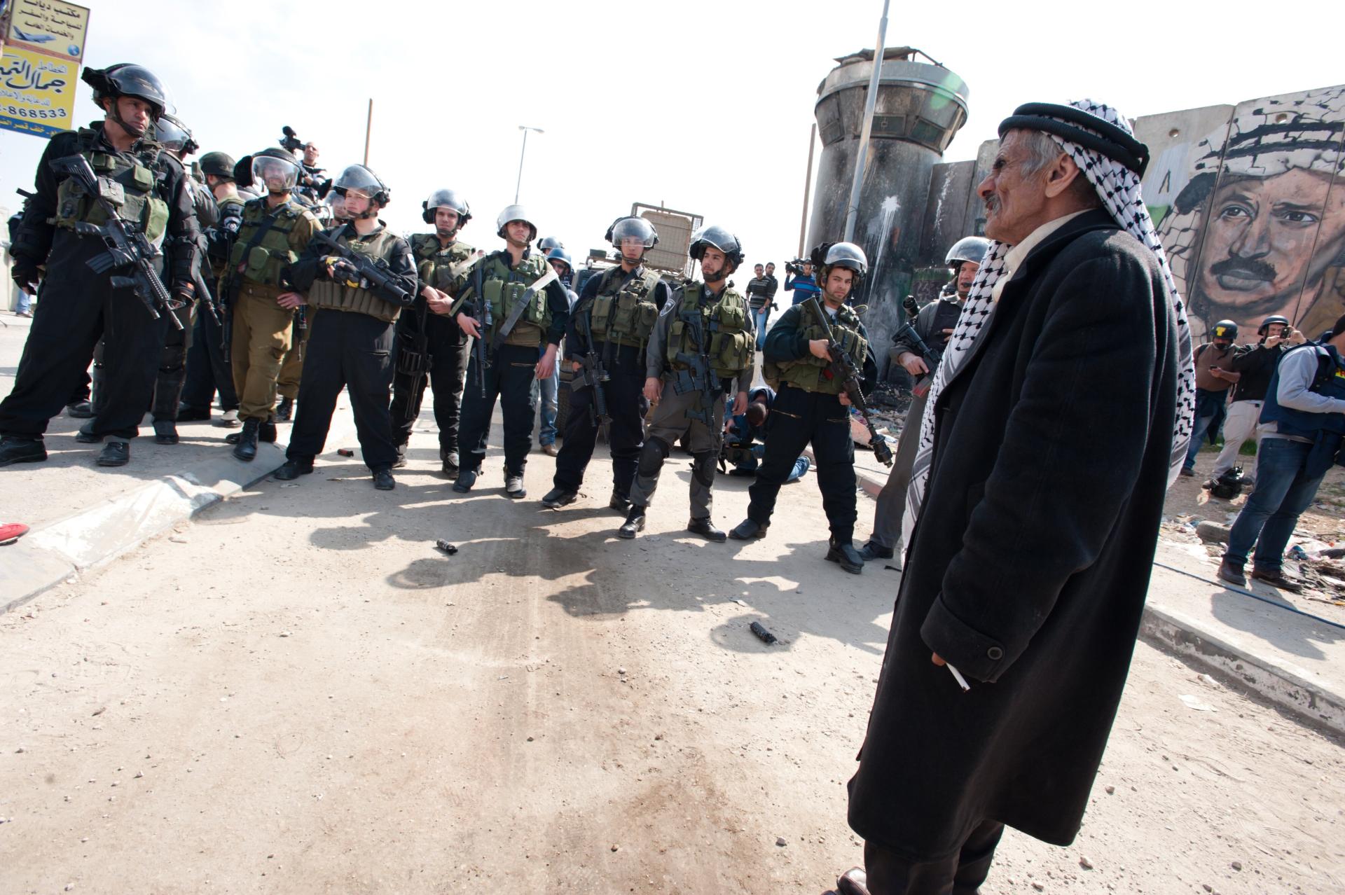 An elderly Palestinian man wearing a traditional 'iqal and kuffiyeh stands near a large group of heavily-armed Israeli soldiers at the Kalandia checkpoint.