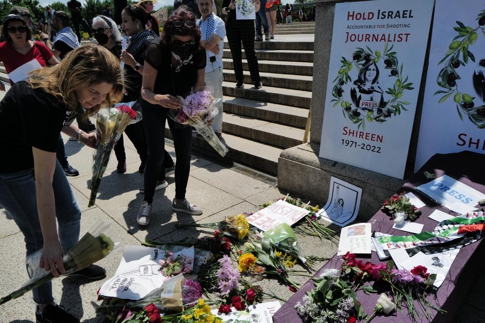 Mourners place flowers at a memorial for Shireen Abu Akleh as part of the Nakba commemoration in Washington DC in 2022, just weeks after she was shot and killed by Israeli forces in Jenin. A poster with her image says "Hold Israel accountable for shooting journalist Shireen, 1971-2022." A symbolic casket is placed on the ground, adorned with flowers, a kuffiyeh, and artwork honoring Abu Akleh.