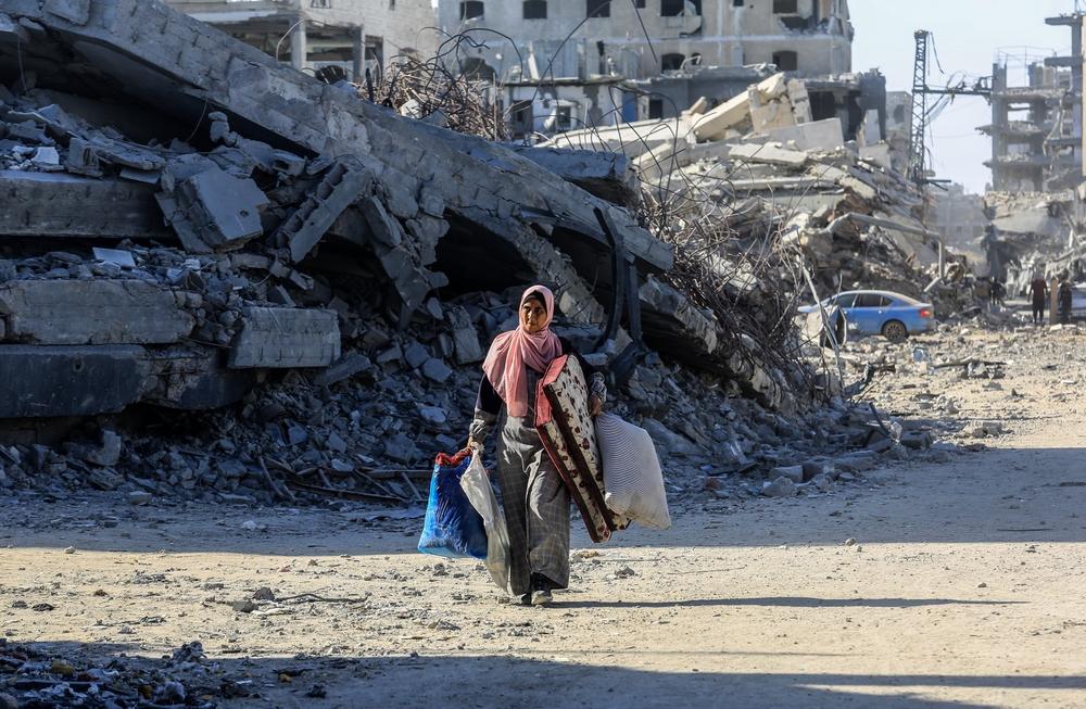 A woman walks near the rubble of a decimated building, carrying bags and large cushions.