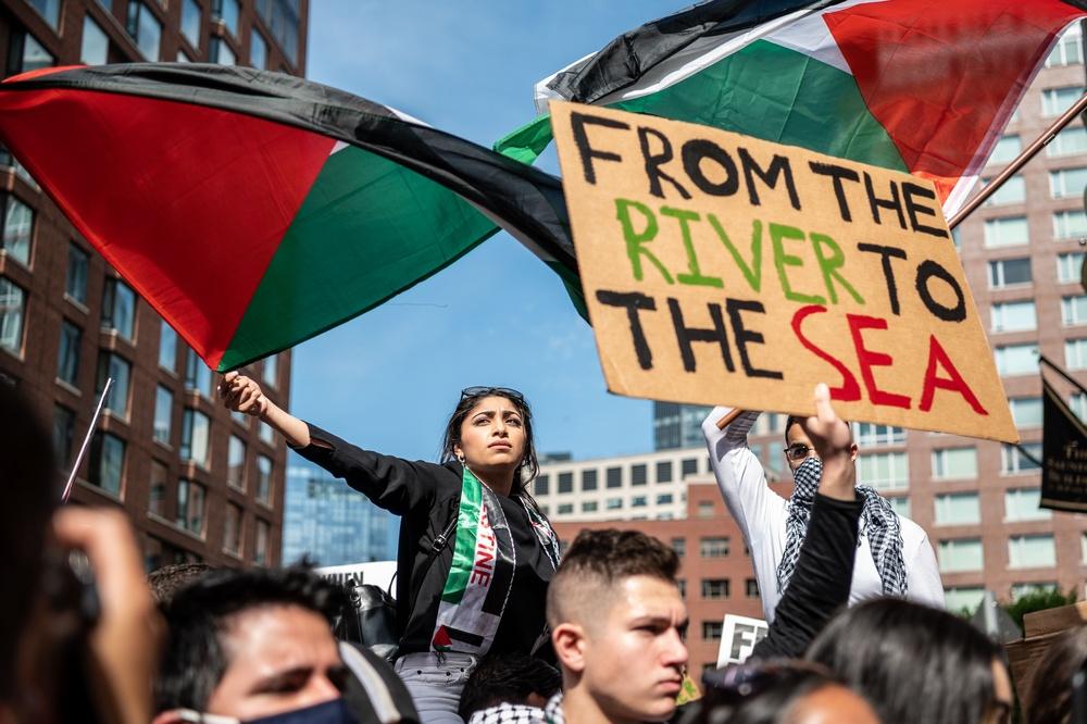 Protest against Israel's 2021 war on Gaza outside of the Israeli Consulate in Boston. Two protestors wave Palestinian flags, one holds a sign that says "FROM THE RIVER TO THE SEA."