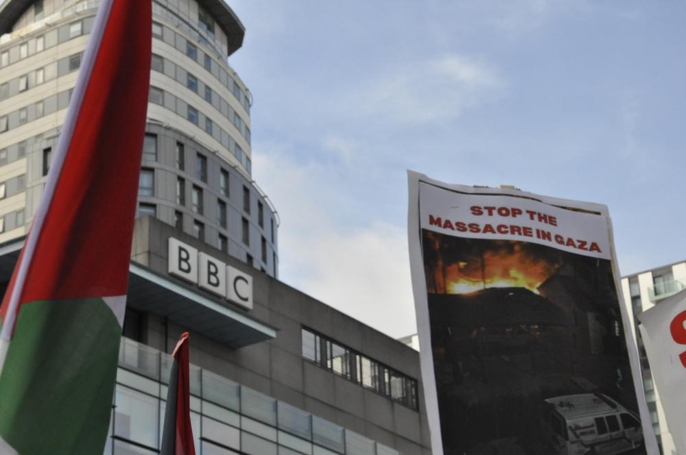 Protest outside of the BBC offices in the UK in October 2023. Image displays BBC name on their building, a Palestinian flag, and a sign that reads "STOP THE MASSACRE IN GAZA" with a widely-circulated image of the Israeli bombing of the Ahli (Baptist) Hospital in Gaza City in October 2023.