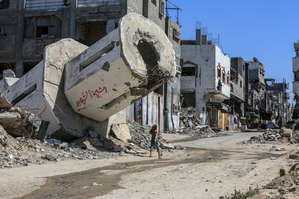 A Palestinian girl walks past a heavily damaged building pillar, hanging over her head, as she walks down a detroyed street in Gaza.