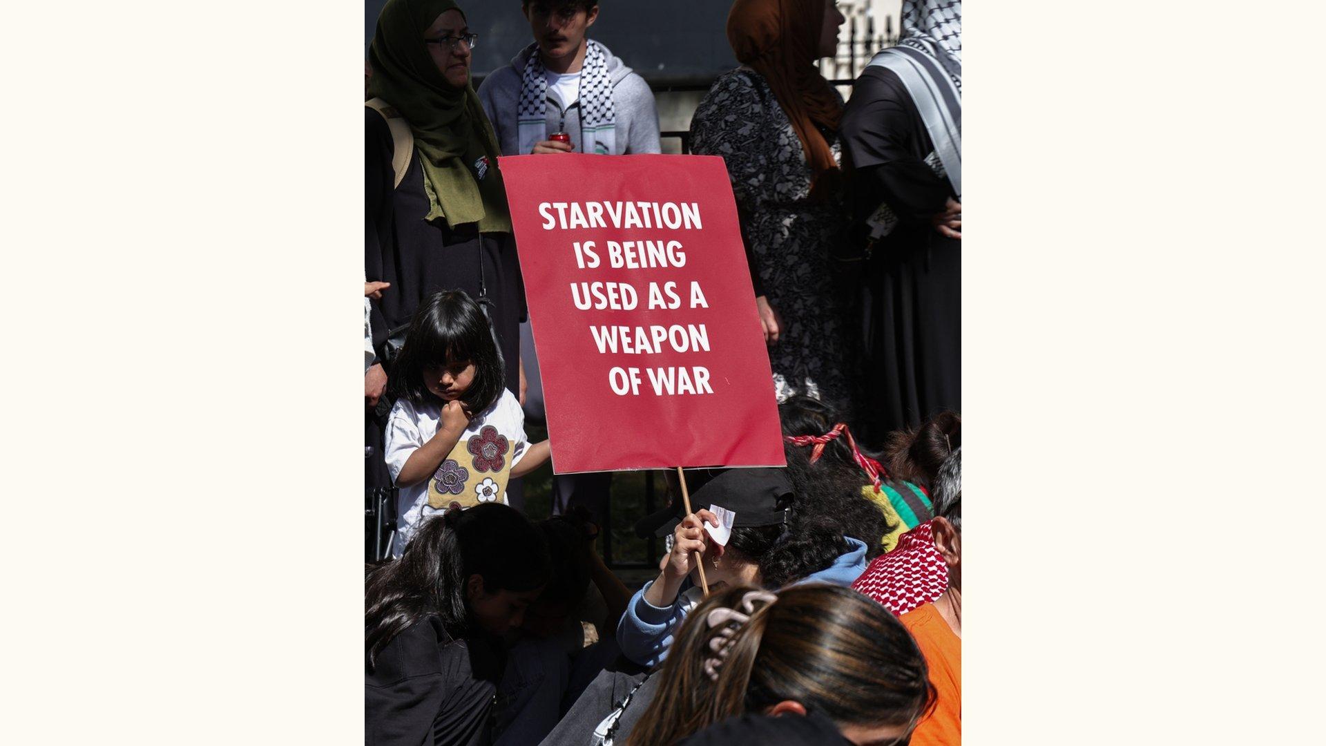 At a protest against Israel's genocide in Gaza, a sign reads "STARVATION IS BEING USED AS A WEAPON OF WAR."