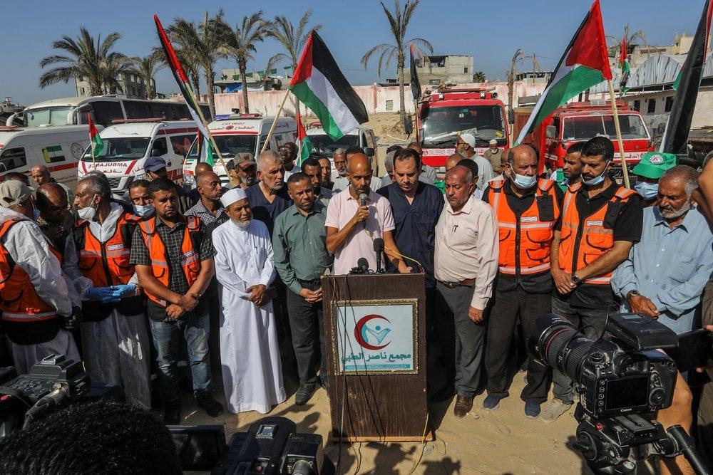 Aid workers, including health professionals and paramedics, participate in a press conference at the Nasser Medical Complex. PRCS ambulances are in the background, some participants hold Palestinian flags.