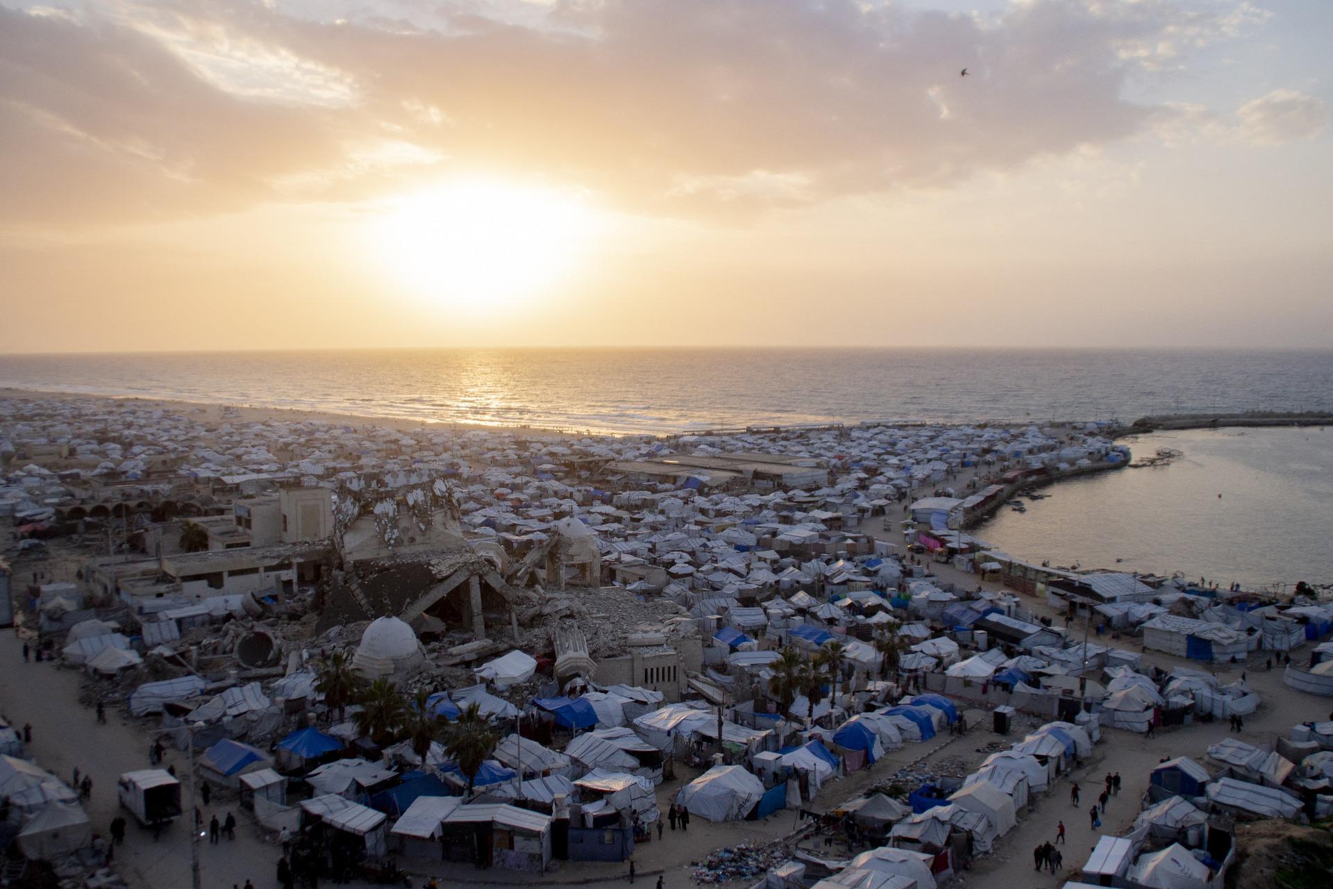Sunset over a crowded makeshift displacement camp along the Gaza seaport, with destroyed rubble of a mosque minaret in view.