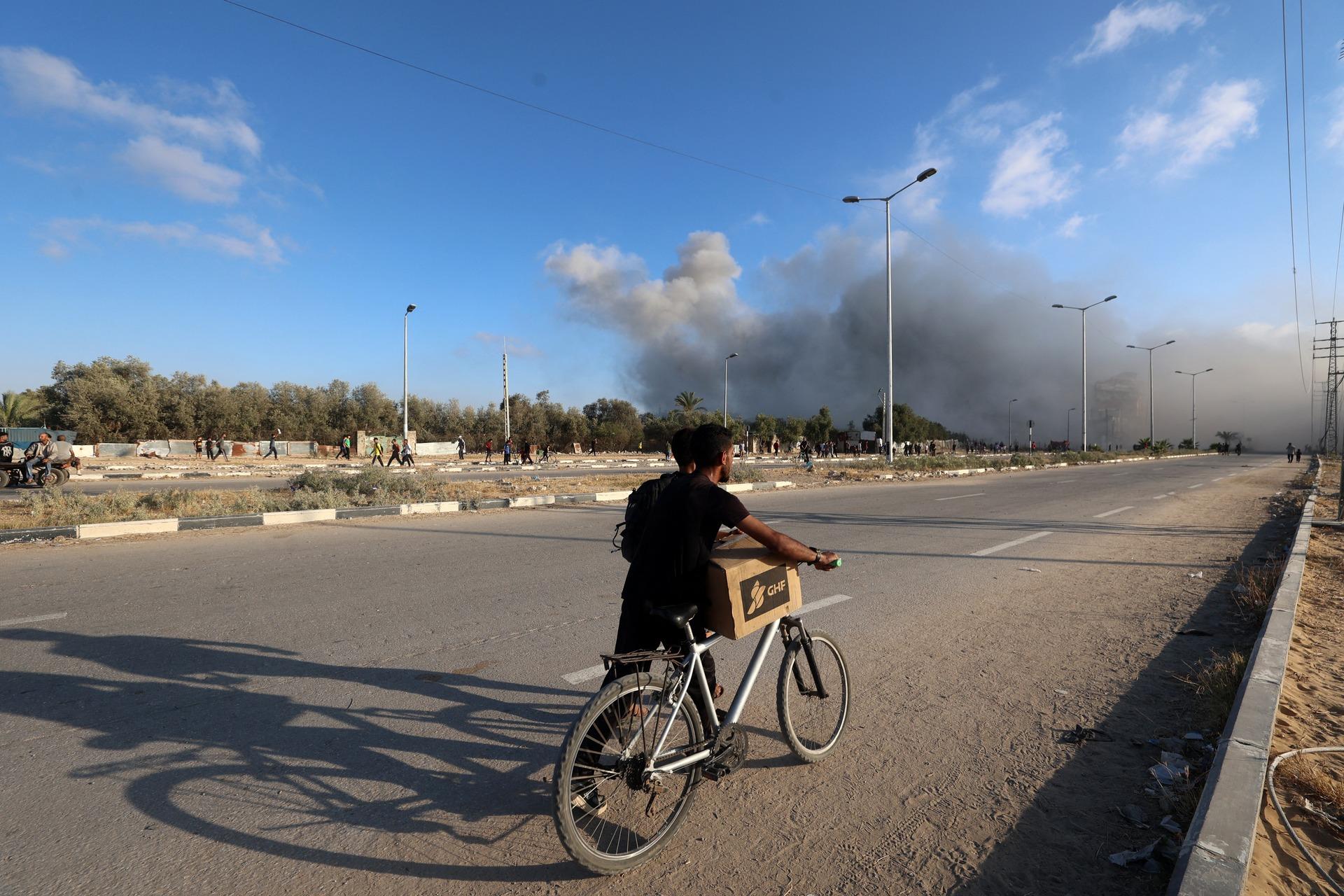A Palestinian walks with a GHF box over his bike seat as smoke rises in the distance along Salah al-Din Road in the central Gaza Strip's Nuseirat refugee camp. June 25, 2025.