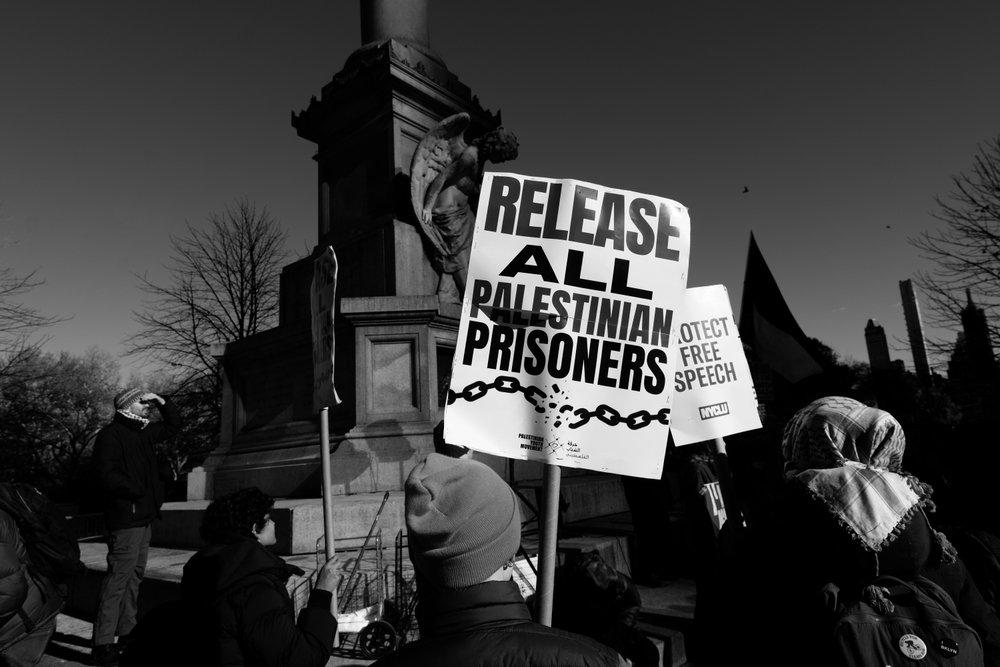 At a Palestine solidarity rally, protestors carry signs that say "RELEASE ALL PALESTINIAN PRISONERS" and "PROTECT FREE SPEECH."