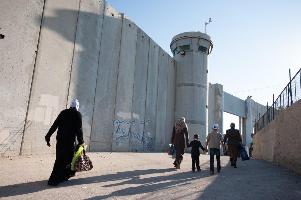 Palestinians walk towards a checkpoint in Bethlehem, with Israel's apartheid wall and a military watchtower in view.
