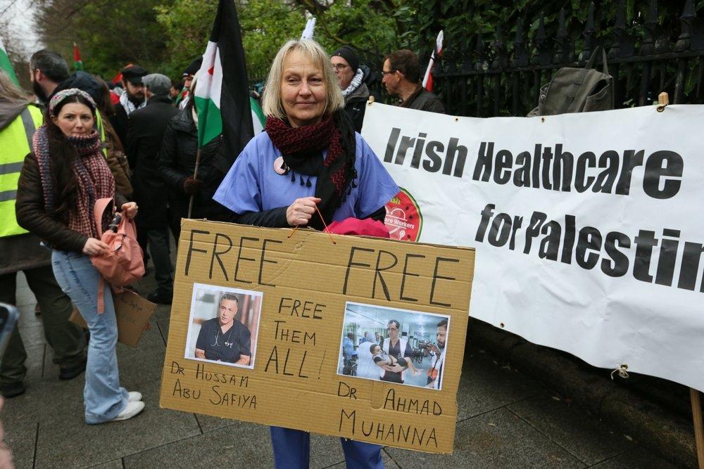At a rally in Dublin, Ireland, a protestor carries a sign with photos of Dr. Hussam Abu Safiya and Dr. Ahmad Muhanna that says "FREE FREE FREE THEM ALL!"