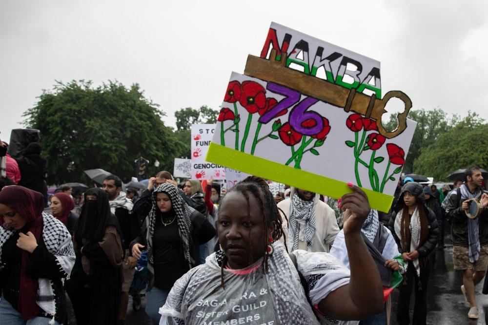 At a rally commemorating the Nakba, a protestor carries a sign that says "NAKBA 78" with a key symbolizing the Palestinian right of return and Palestinian poppy flowers.