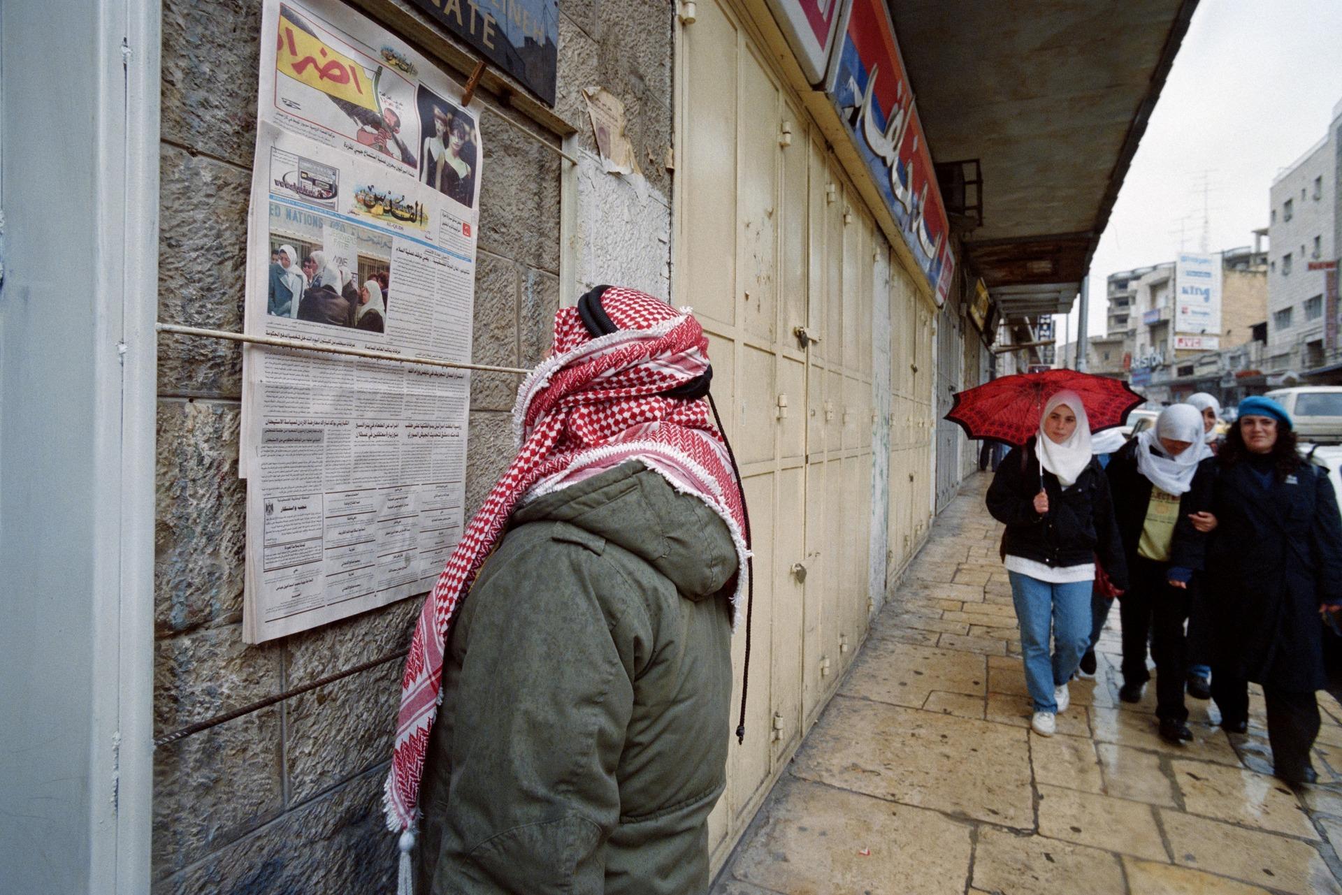 In Occupied East Jerusalem, a Palestinian man reads an article in Al-Quds newspaper on the strike in protest of Israel settlement expansion. A group of young women walks by. Shops are closed as part of the general strike. March 3, 1997.