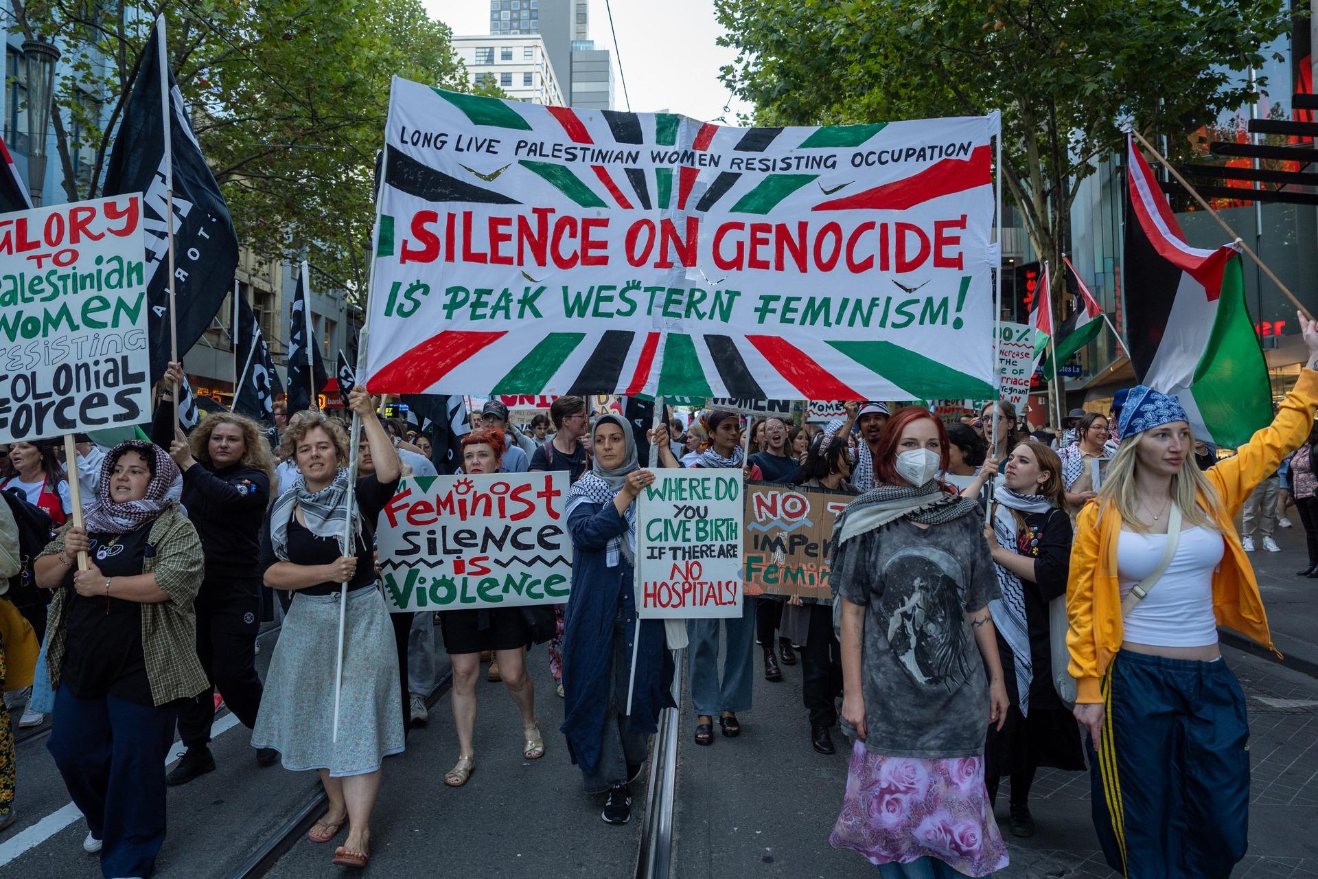 Protestors in Melborne, Australia, hold up signs that say "Where do you give birth if there are no hospitals," "feminist silence is violence," and "LONG LIVE PALESTINIAN WOMEN RESISTING OCCUPATION: SILENCE ON GENOCIDE IS PEAK WESTERN FEMINISM!"