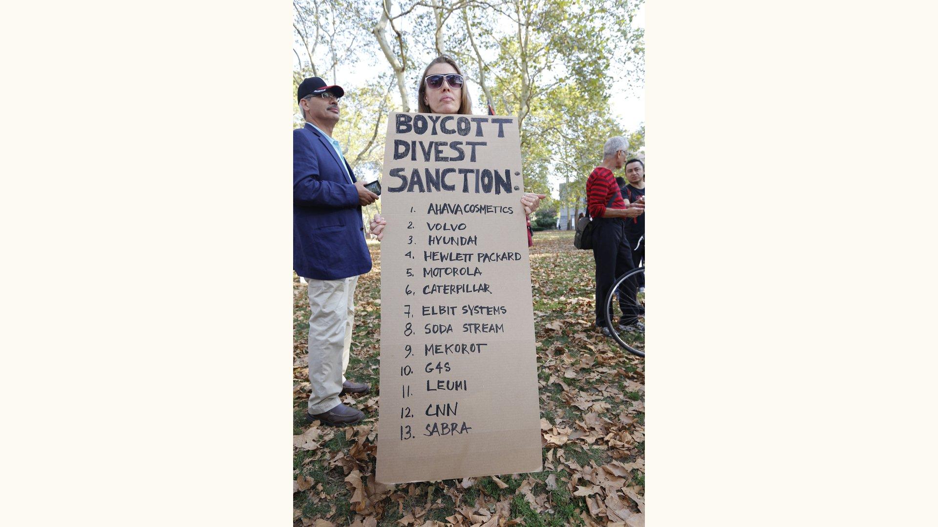 A protest sign reads "BOYCOTT DIVEST SANCTION: AHAVA COSMETICS, VOLVO, HYUNDAI, HP, MOTOROLA, ELBIT, SODASTREAM, MEKOROT, G4S, LEUMI, CNN, SABRA"