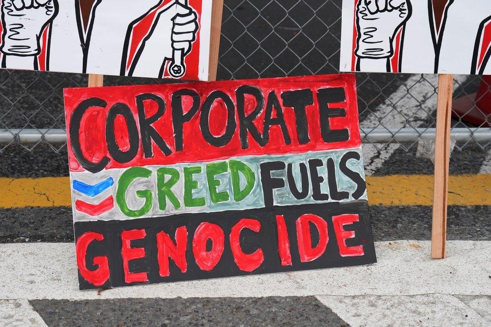 At a protest at the Chevron refinery in Richmond, CA a sign reads "CORPORATE GREED FUELS GENOCIDE" beside the Chevron logo.