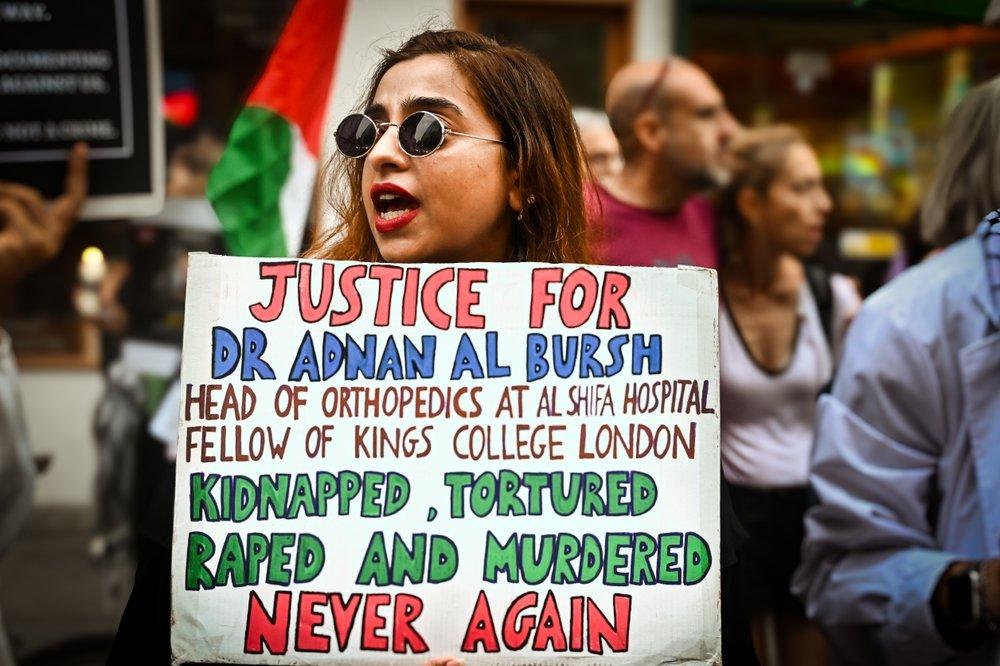 A protestor carries a sign that reads "JUSTICE FOR DR. ADNAN AL BURSH, HEAD OF ORTHOPEDICS AT AL-SHIFA HOSPITAL, FELLOW OF KINGS COLLEGE LONDON: KIDNAPPED, TORTURED, RAPED AND MURDERED.. NEVER AGAIN."