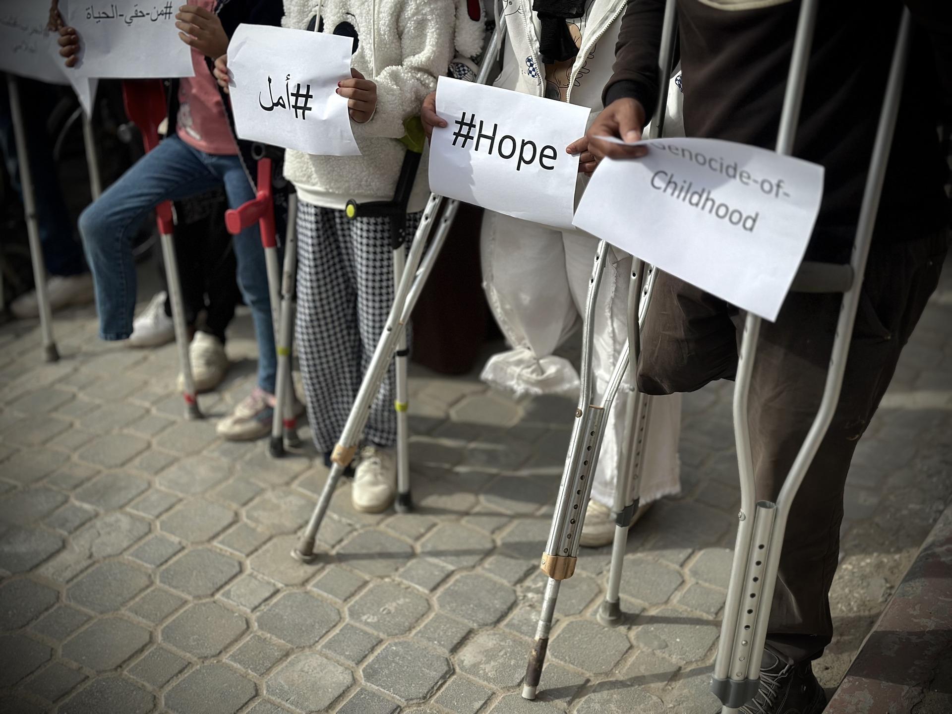 Palestinian children who were forced to have limbs amputated due to Israel's genocidal war on Gaza appear at a rally in front of Nasser Hospital in Gaza. Image focuses on their crutches and amputated limbs, and the protest signs they carry, which read "genocide of childhood," and "hope."