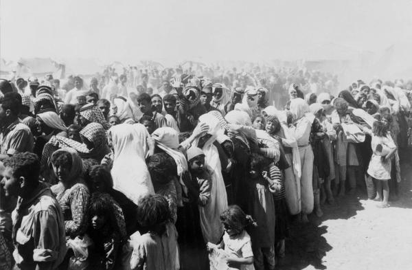 A large crowd of Palestinian refugees line up at the Wadi Dalail Refugee Camp in Jordan under heavy dust, with their tents in the background.