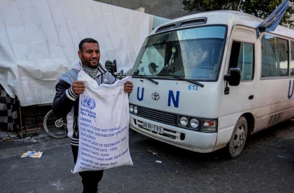 A Palestinian carries a bag of UNRWA flour, distributed by the agency to refugees in need of humanitarian aid, with a marked UN van bearing the UN flag in the background.