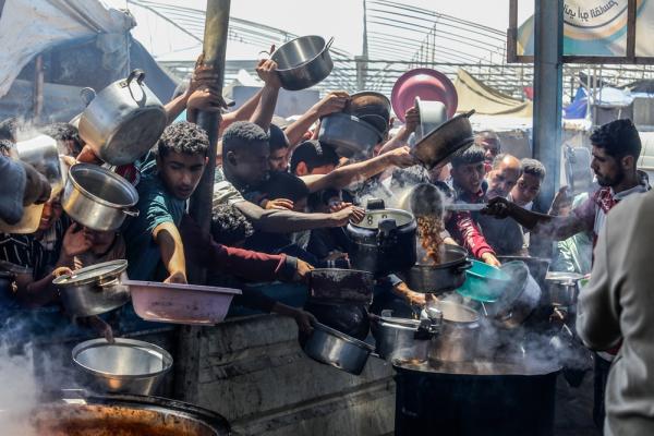 A large crowd of Palestinians carry pots and large bowls as aid workers distribute cooked meals in Mawasi, Khan Yunis as Israel's forced starvation of the Gaza Strip intensifies. April 2025.
