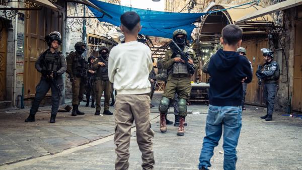 Two Palestinian boys look on as heavily armed Israeli soldiers block access to a street in Hebron's old city. One masked soldier wearing dark sunglasses and carrying a weapon walks towards the boys.