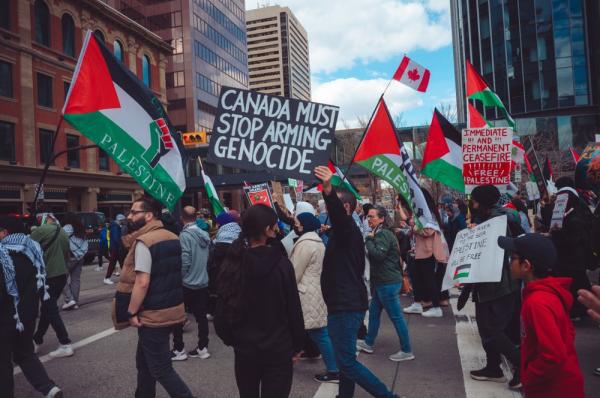Protestors in Calgary, Canada wave Palestinian flags, a Canadian flag, and signs demanding a ceasefire in Gaza. One prominent sign reads "CANADA MUST STOP ARMING GENOCIDE."