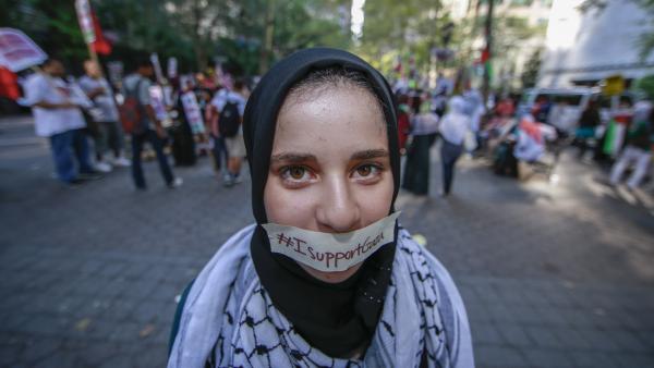 A protestor at a rally against Israel's 2014 war on Gaza in New York City, wearing a kuffiyeh, has tape over her mouth with the words "#ISupportGaza," symbolizing efforts to silence free speech on Palestine.