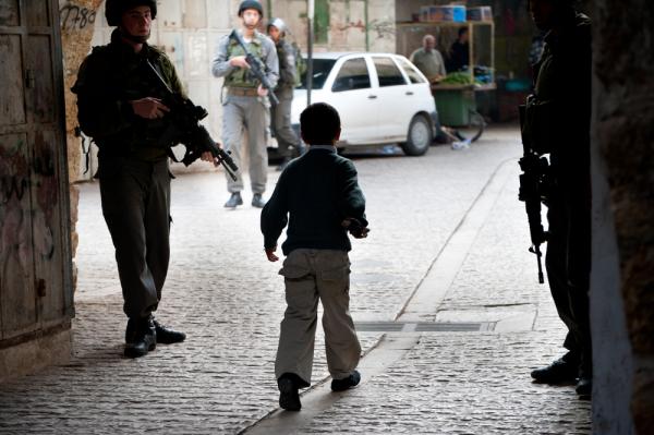 A Palestinian boy walks on a Hebron street surrounded by three Israeli soldiers carrying arms.