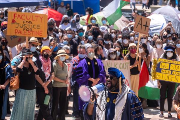 A speaker wearing a graduation cap and gown, and a kuffiyeh, addresses the anti-genocide encampment at UCLA. Members of the large crowd carry signs, one says "FACULTY FOR JUSTICE FOR PALESTINE."