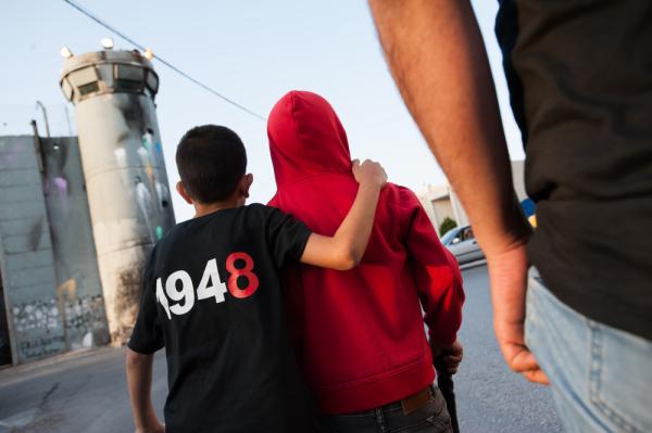 A Palestinian boy walks near Israel's apartheid wall and a watchtower with an arm around his friend's shoulder. The back of his shirt reads "1948." 