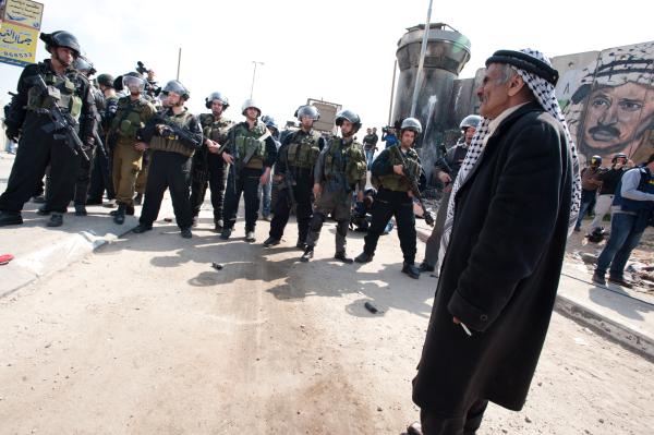 An elderly Palestinian man wearing a traditional 'iqal and kuffiyeh stands near a large group of heavily-armed Israeli soldiers at the Kalandia checkpoint.