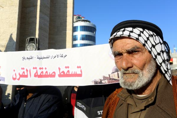 A Palestinian elder wearing a traditional 'iqal and kuffiyeh carries an Arabic sign that says "down with the deal of the century."