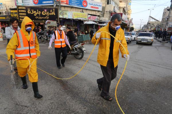 Municipal workers wearing masks and Personal Protective Equipment (PPE) carry a high-pressure power sprayer for deep cleaning and mass-sanitization in the earliest days of the COVID-19 pandemic. Rafah, Gaza Strip. March 14, 2020.