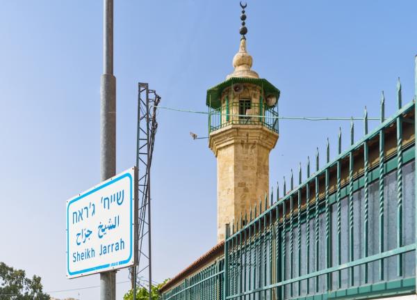 Street sign marking the Sheikh Jarrah neighborhood in Arabic, English, and Hebrew. Behind a fence is a minaret.