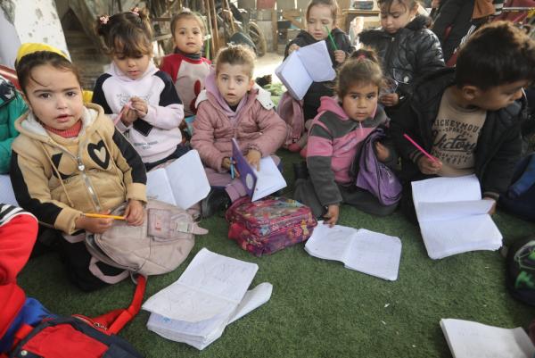 Displaced Palestinian schoolchildren sit on the floor of a classroom in Khan Yunis partially destroyed in Israel's genocide of Gaza with notebooks, backpacks, and pencils.