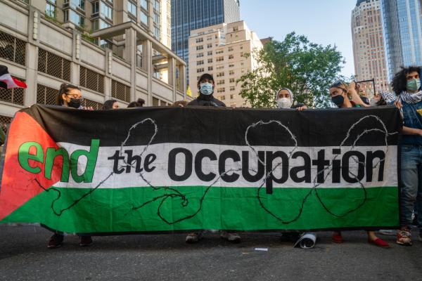 Protestors carry banner that says "end the occupation" over a Palestinian flag in the background, with barbed wire running over the banner.