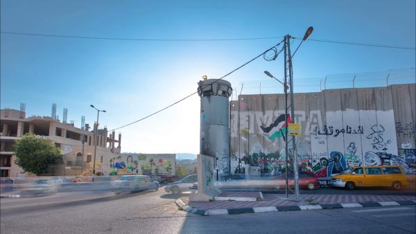 View of Israel's apartheid wall and a watchtower cutting through the West Bank city of Bethlehem as carys drive by. Graffiti on the apartheid wall features a Palestinian flag and other Palestinian imagery.
