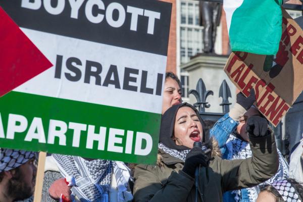Protestors at a rally in Boston  wear kuffiyehs and carry signs that say "Boycott Israeli Apartheid" over a Palestinian flag and "Free Palestine."