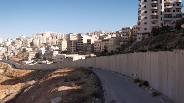 View of Israel's apartheid wall, separating a road and hilly landscape from an area with many buildings.